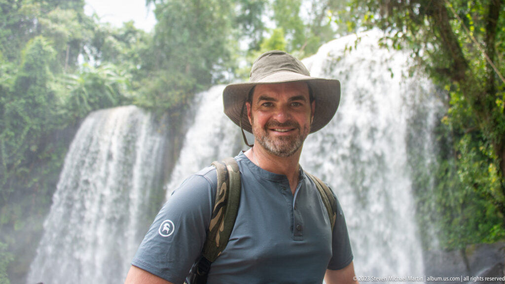 Man in a sun hat and backpack stands smiling before a broad waterfall.