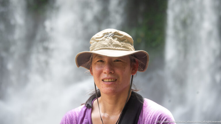 Woman in a beige sun hat and purple shirt poses with waterfall spray behind.