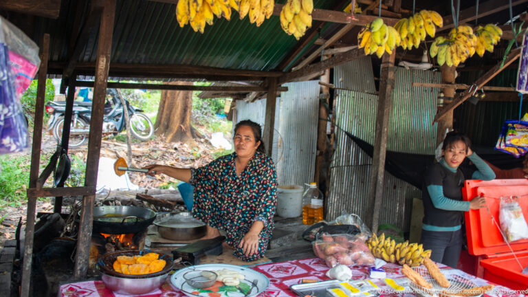 Two women prepare food in a rustic stall with hanging bananas, pots, and a wood fire.