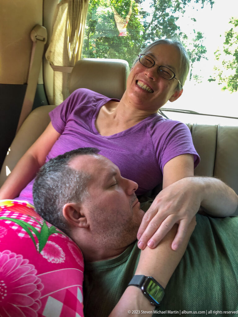 Smiling woman supports a reclining man in the back seat beside a floral cushion.