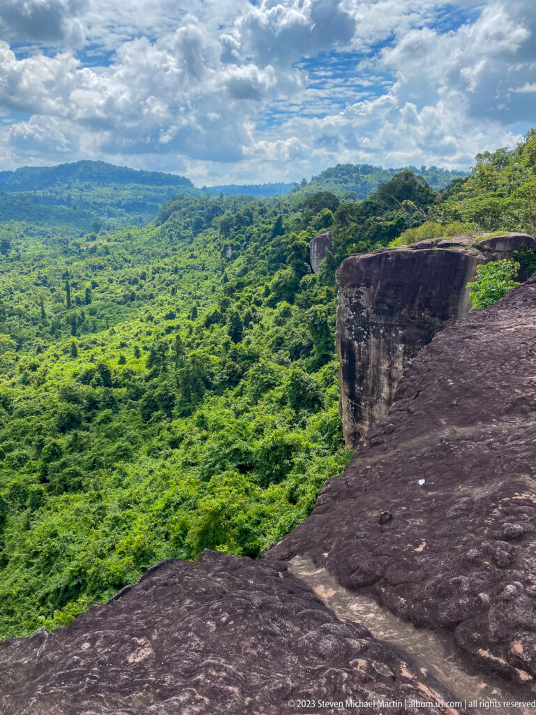 Dark rock shelf and cliff edge overlook an expansive sunlit green forest plateau.