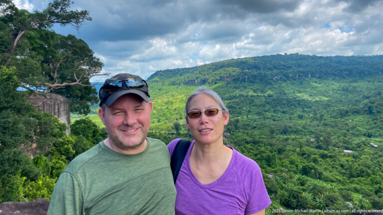 Two adult visitors pose together above a green valley and forested hills.
