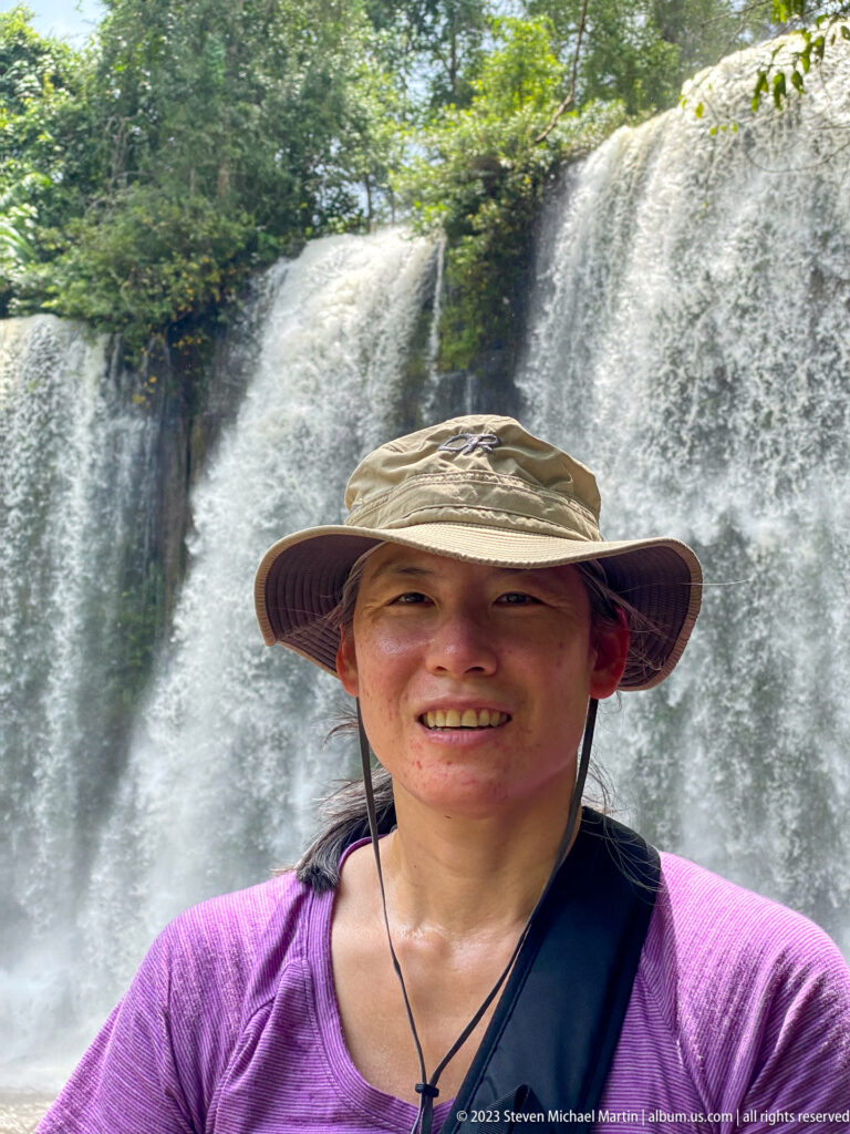 Visitor in a sun hat smiling with a waterfall behind.