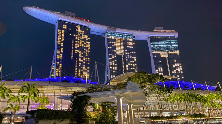 Marina Bay Sands at night above blue-lit canopies, palms, and waterfront structures.