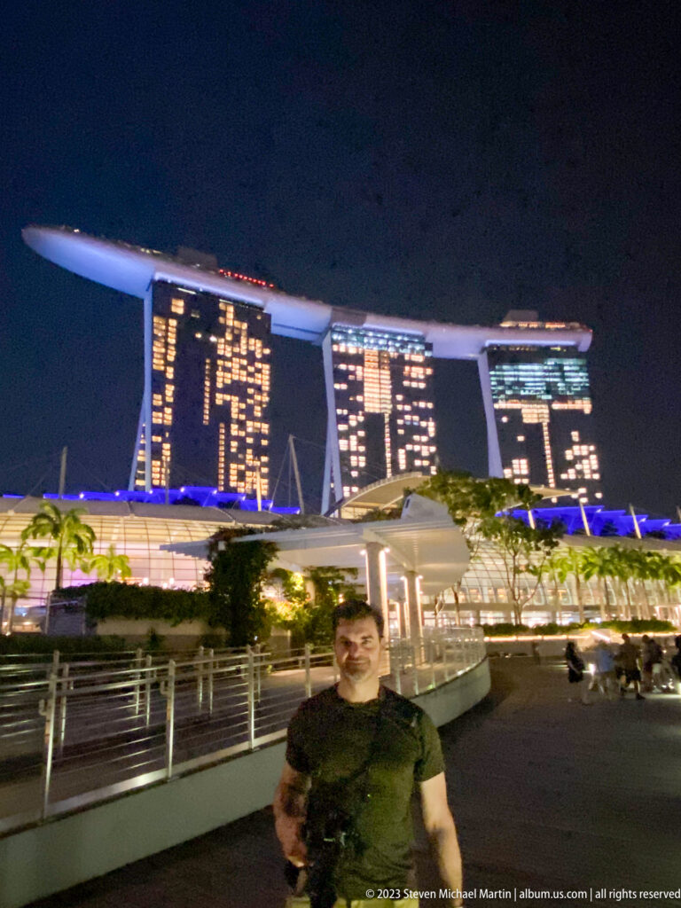 Man standing on a waterfront promenade with Marina Bay Sands illuminated behind him.