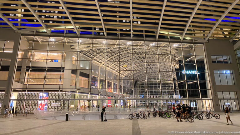 Large glass entrance to The Shoppes at Marina Bay Sands with people and bicycles in front.