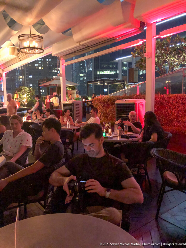 People seated at a red-lit outdoor bar terrace with city towers visible behind.