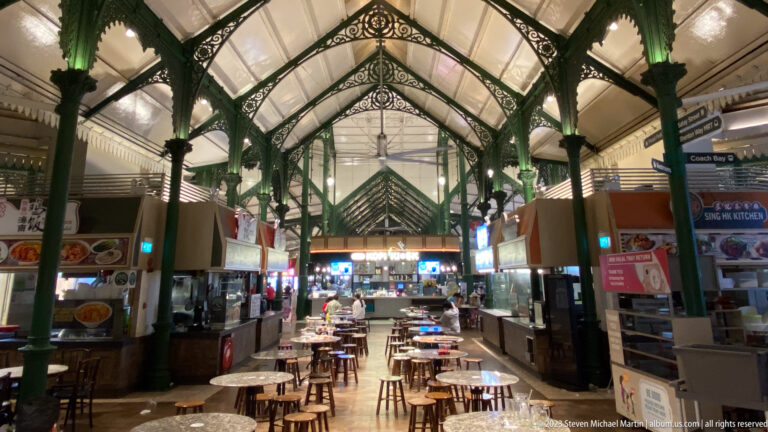 Interior of Lau Pa Sat with green iron arches, food stalls, and mostly empty tables.