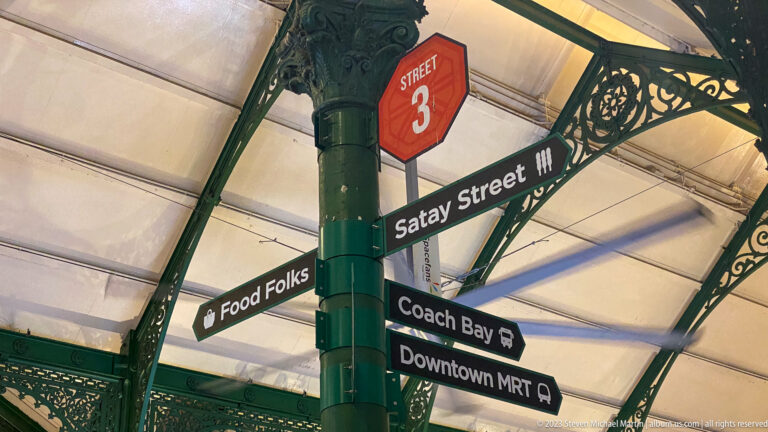 Directional signs for Satay Street, Food Folks, Coach Bay, and Downtown MRT on a green iron column.