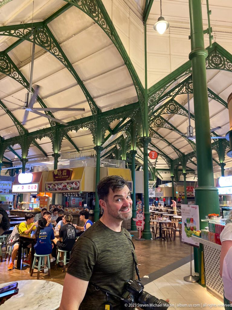 Man standing inside Lau Pa Sat with food stalls, diners, and green iron columns behind him.