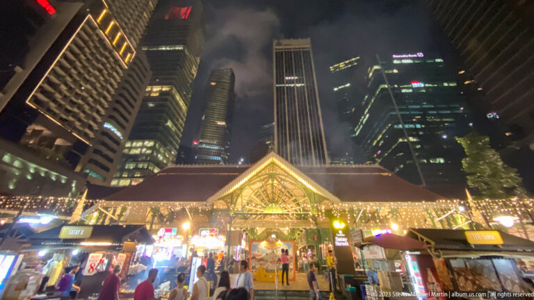Illuminated Lau Pa Sat entrance with food stalls, visitors, and tall office towers behind.