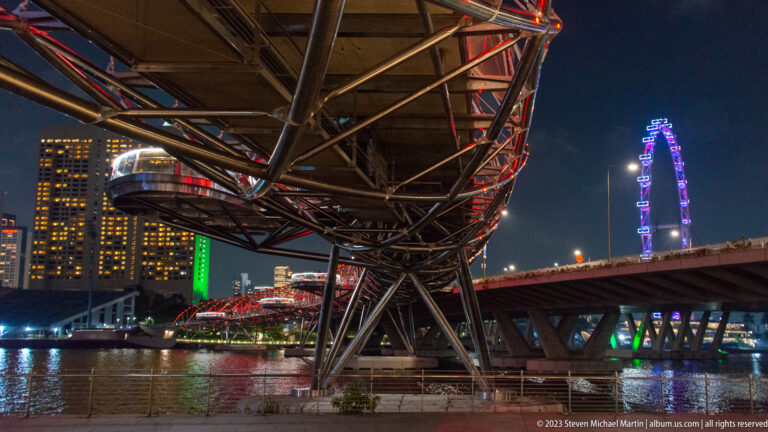 Underside of the red-lit Helix Bridge with a viewing pod and Singapore Flyer in the background.