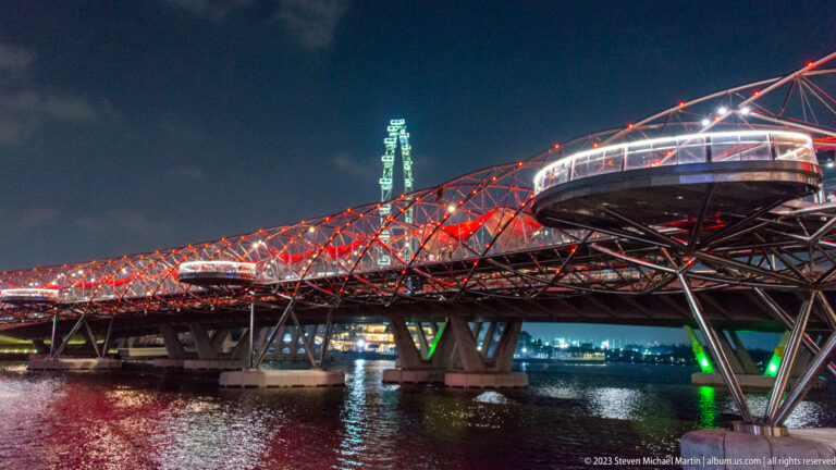 Side view of the red-and-white illuminated Helix Bridge with viewing pods over Marina Bay.