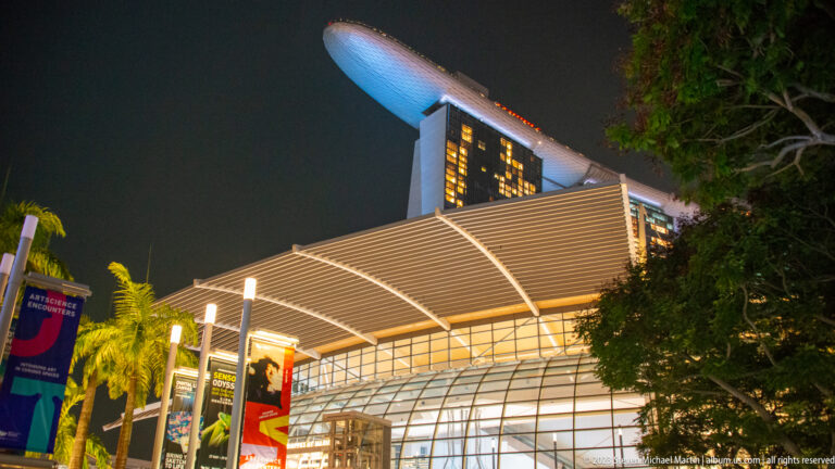 Marina Bay Sands visible above a bright ribbed canopy, trees, and waterfront banners at night.