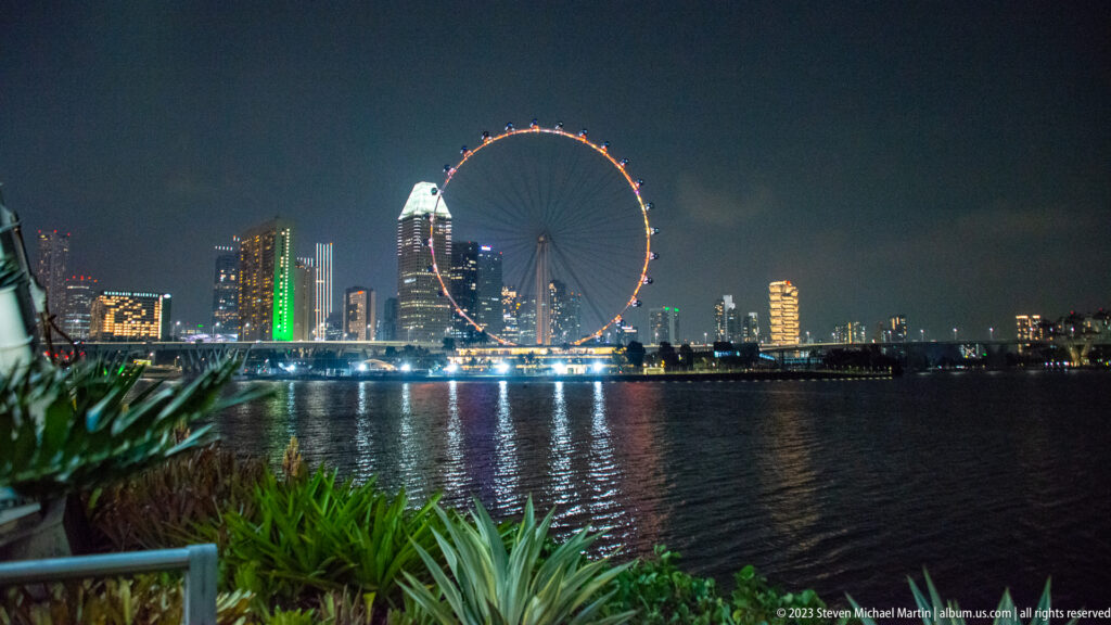 Singapore Flyer and illuminated skyline across Marina Bay, with plants and railing in the foreground.