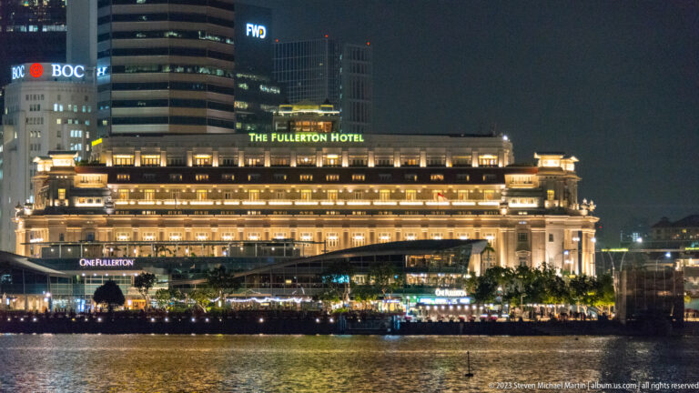 The illuminated Fullerton Hotel across the water with modern office towers behind it.