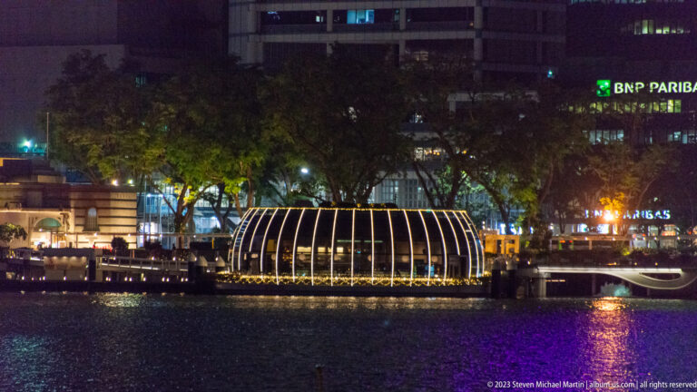 Rounded ribbed glass pavilion glowing across Marina Bay with purple reflections and office towers behind.