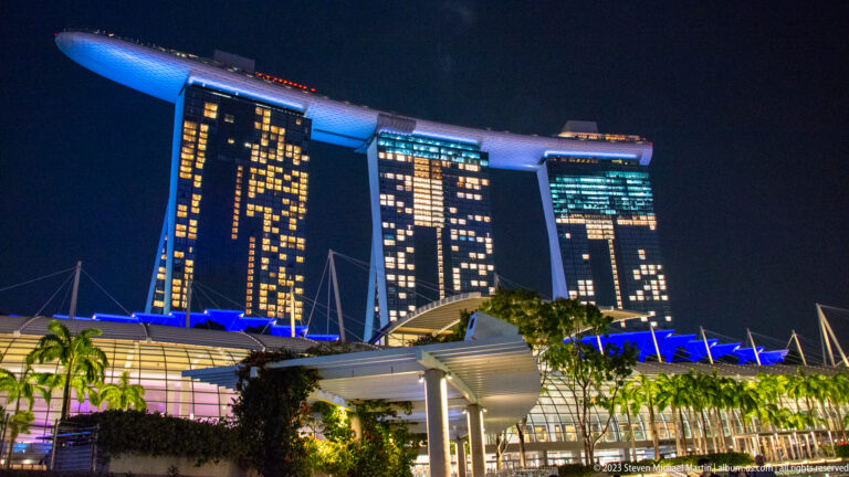 Marina Bay Sands at night above palms, a bright canopy, and waterfront structures.