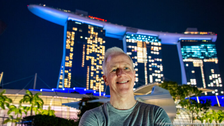 Smiling gray-haired man in front of the illuminated Marina Bay Sands hotel at night.