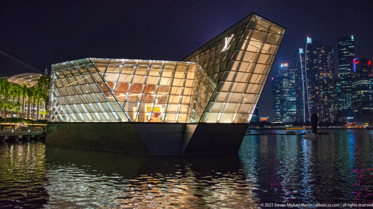 Illuminated faceted Louis Vuitton pavilion on Marina Bay with downtown towers reflected behind it.