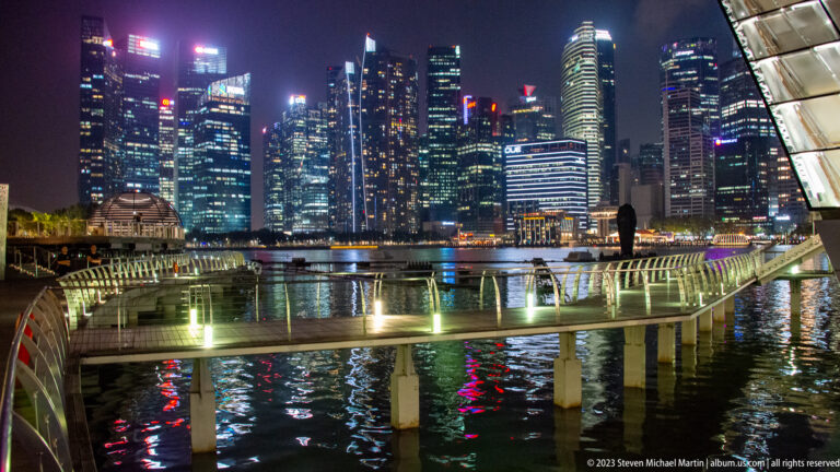 Curving lit walkway over Marina Bay with the illuminated downtown Singapore skyline beyond.