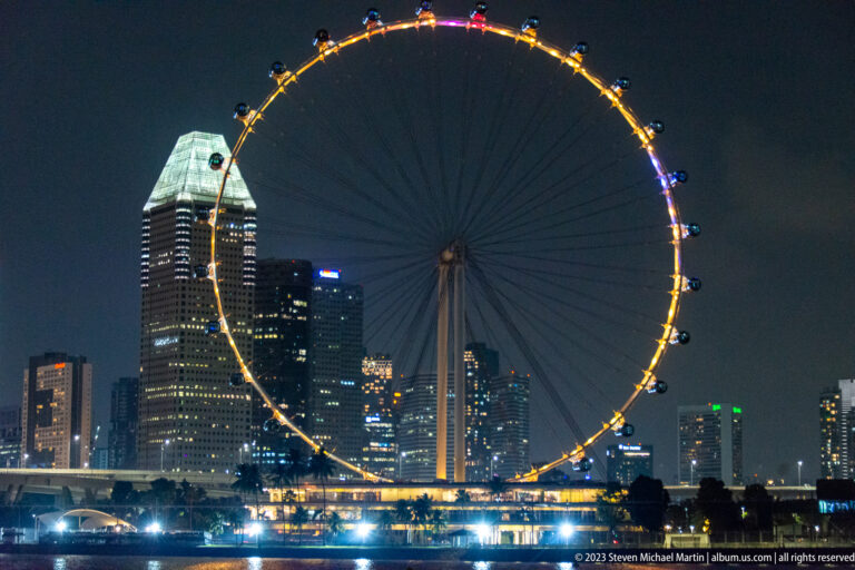 Close nighttime view of the Singapore Flyer with skyscrapers and waterfront lights behind it.