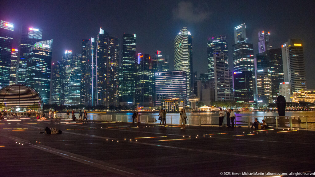 People on a lit Marina Bay promenade facing Singapore’s illuminated downtown skyline across the water.