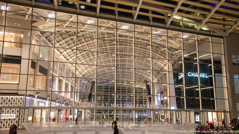 Large glass entrance to The Shoppes at Marina Bay Sands with illuminated retail levels and brand signs.