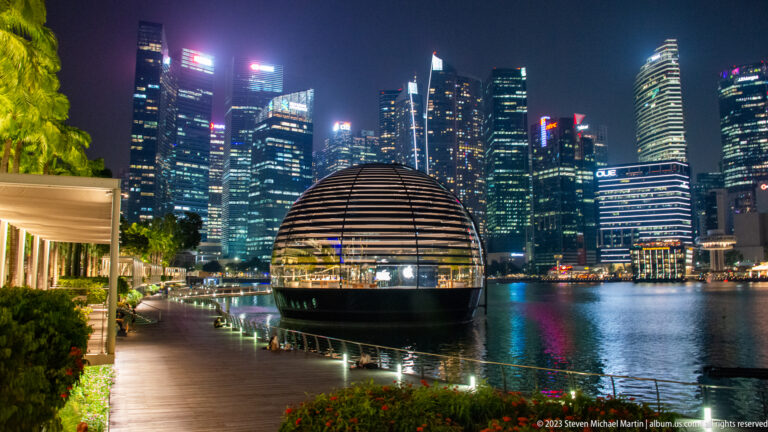 Round banded Apple pavilion beside Marina Bay with illuminated skyscrapers behind it.