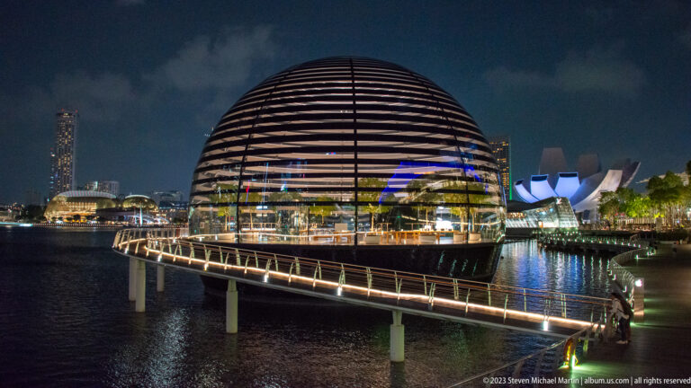 Lit curved footbridge leading to the round Apple pavilion over Marina Bay at night.