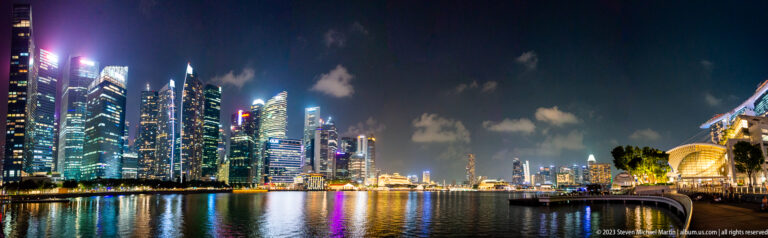 Wide nighttime panorama of Marina Bay with downtown skyscrapers and waterfront buildings reflected in the water.