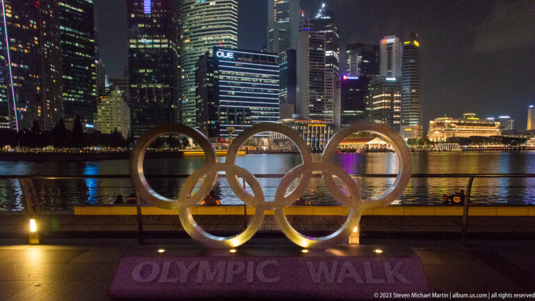 Illuminated Olympic rings sculpture on the waterfront with Singapore’s skyline across Marina Bay.