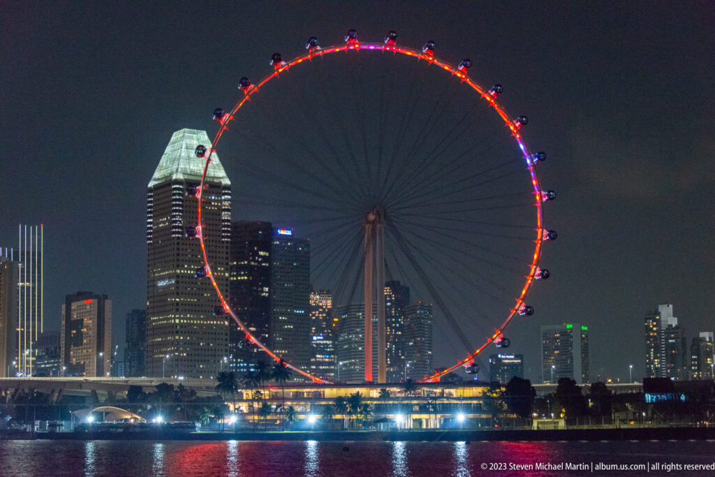 Singapore Flyer glowing red above waterfront buildings, with colored reflections on Marina Bay.