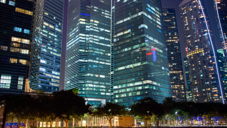 Closely grouped illuminated glass office towers rising above trees in central Singapore.
