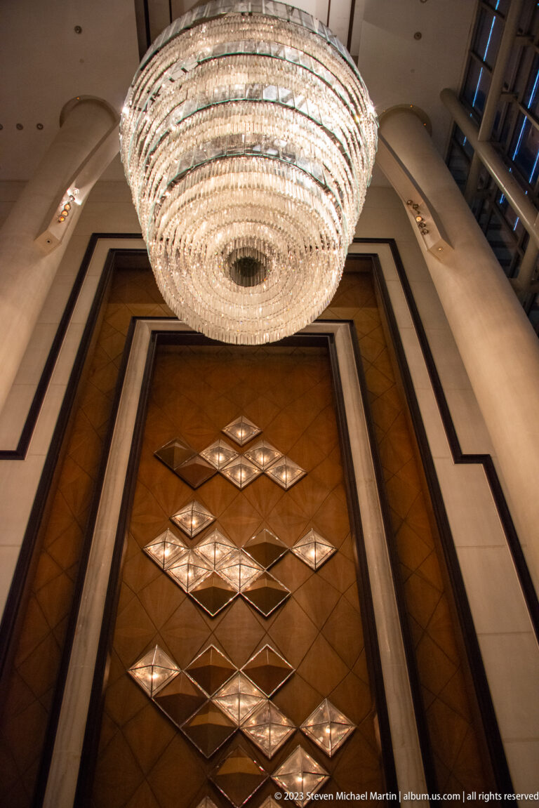 Large circular crystal chandelier above a tall wood-paneled wall with glowing diamond patterns.