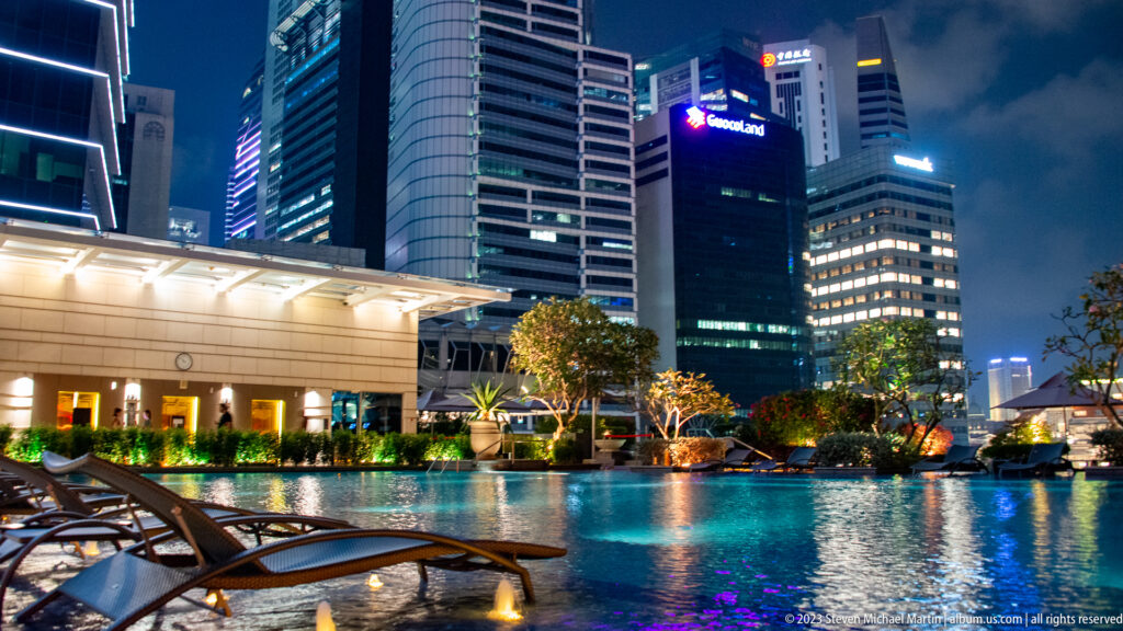 Rooftop pool with lounge chairs and landscaped edges beneath illuminated Singapore office towers.