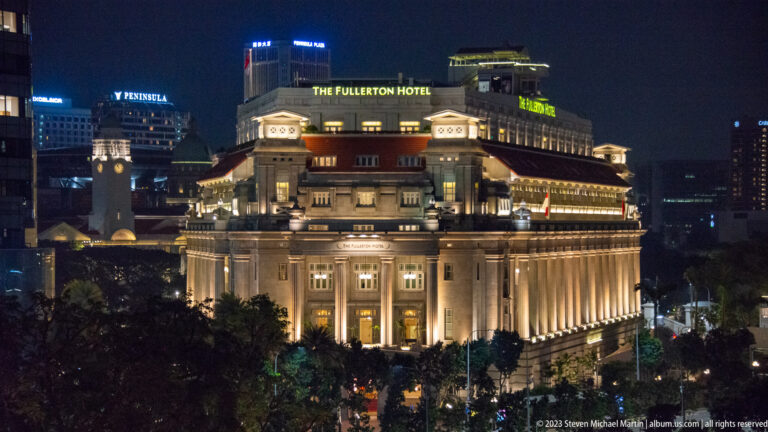 Illuminated Fullerton Hotel viewed from above, surrounded by trees and city towers at night.