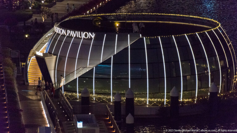 Rounded glass pavilion with vertical light strips beside a dark waterfront walkway.