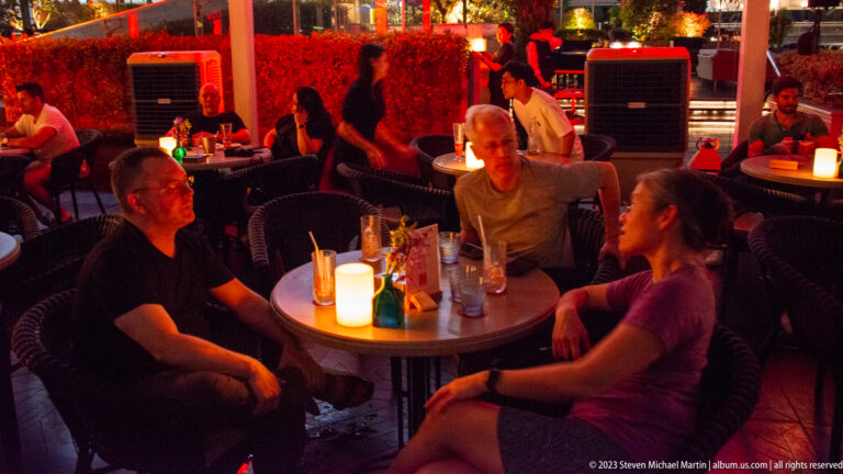 Three adults talking around a candlelit table at a busy outdoor bar at night.