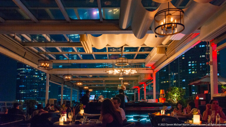 Open-air restaurant with seated diners, hanging lanterns, and illuminated city towers outside.
