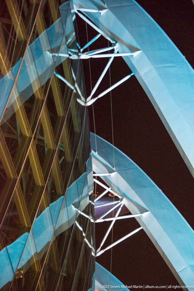 Close view of illuminated white steel beams crossing beside reflective blue and amber glass panels.