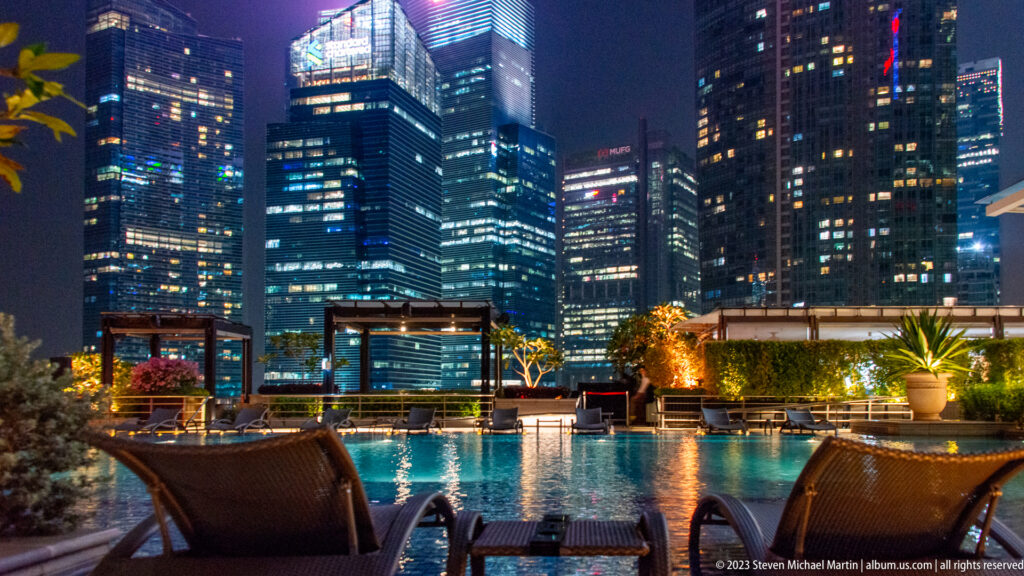 Two lounge chairs beside a rooftop pool with illuminated office towers beyond.