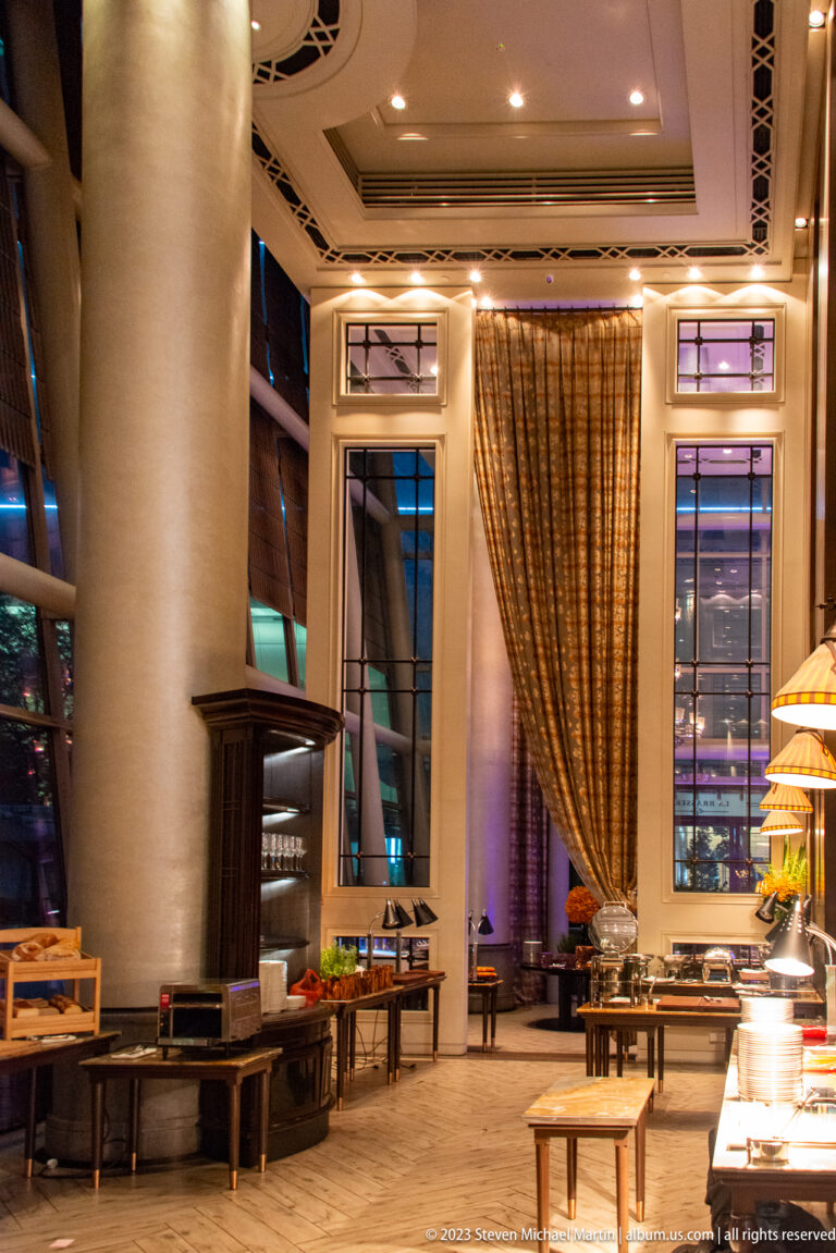 Elegant hotel dining room with tall columns, draped windows, buffet stations, and warm lighting.