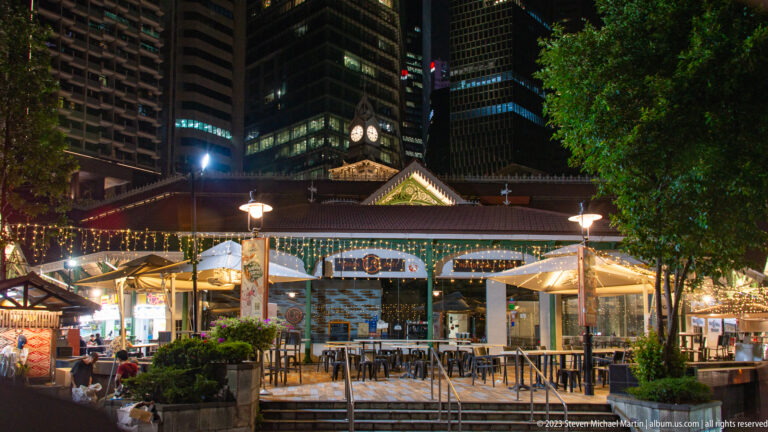 Illuminated Lau Pa Sat market with outdoor tables, umbrellas, clock tower, and office buildings behind.