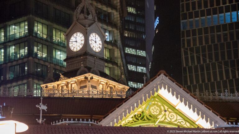 Close view of Lau Pa Sat’s illuminated clock tower and ornate roof gables beneath modern towers.