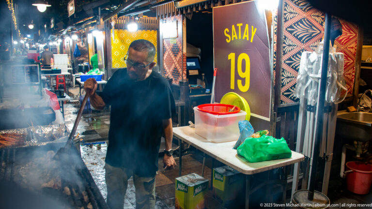 Man grilling satay skewers at a smoky hawker stall marked Satay 19.