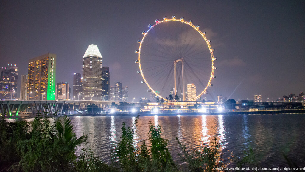 Golden Singapore Flyer across Marina Bay with shrubs in front and a bright reflection on the water.