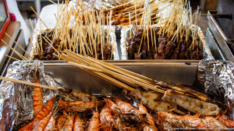 Foil-lined hawker grill filled with satay skewers, prawns, and other grilled seafood.
