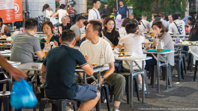 Busy hawker-centre seating area with groups eating and talking at closely spaced tables.