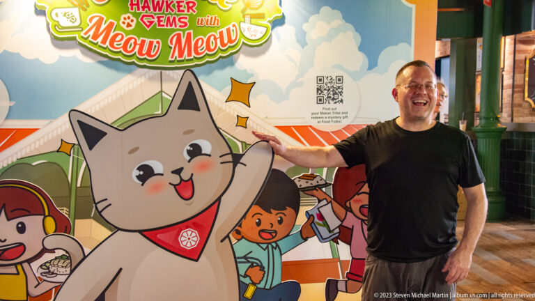 Smiling man pointing toward a large cartoon cat on a colorful hawker-themed mural.